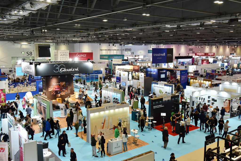 Overhead view of a busy beauty trade show exhibition hall with branded booths and crowds at the Professional Beauty London