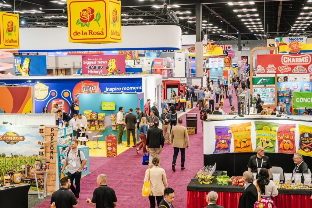 Busy Sweets & Snacks Expo trade show floor, where attendees browse colorful confectionery and snack booths