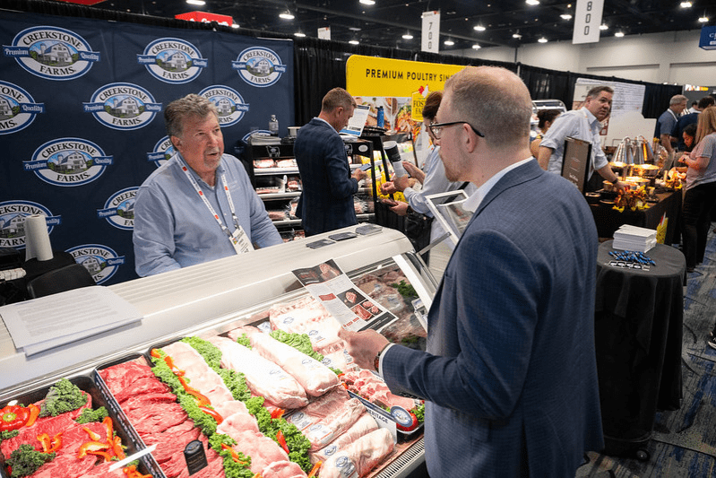 Exhibitors and attendees discussing meat products at a trade show booth during The Annual Meat Conference