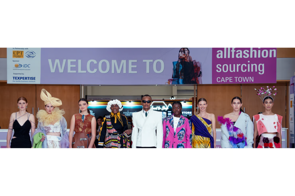 Models in diverse fashion designs stand before an allfashion sourcing Cape Town welcome banner.